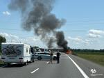 płonący samochód na autostradzie, ogień i kłęby dymu, radiowóz zabezpiecza przejazd na drodze, pasażerowie samochodów stoją na drodze